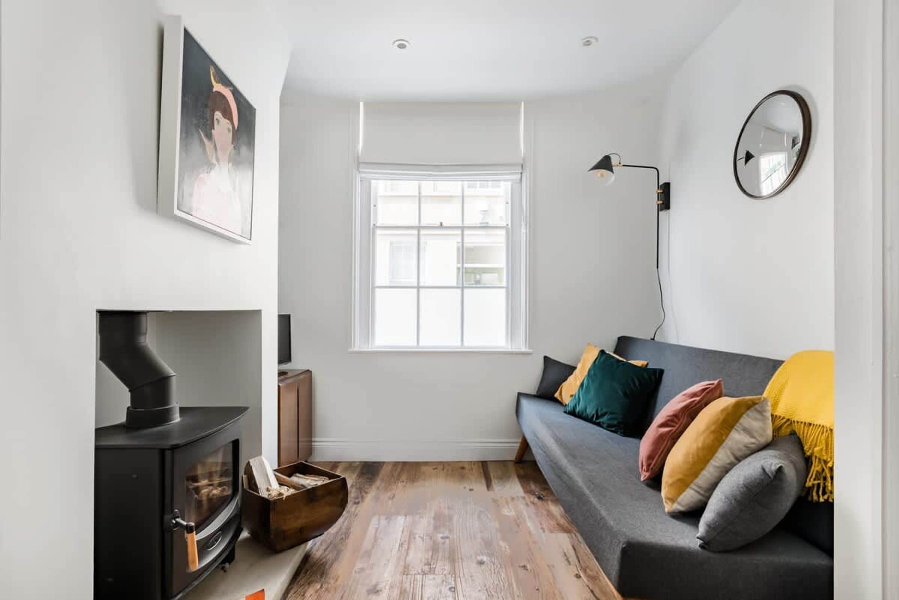 Ein modernes Wohnzimmer mit einem schwarzen Kaminofen, einem grauen Sofa mit bunten Kissen, einem großen Fenster und einem wandmontierten Spiegel.