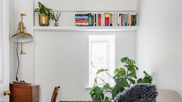 Ein gemütliches Zimmer mit Holzboden, einem bequemen Ohrensessel, einem kleinen Holztisch, einer Heizung, einem Bücherregal mit Büchern und einem Fenster mit Pflanzen.