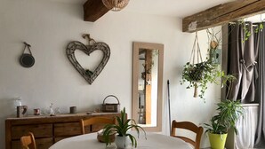 Un comedor con una mesa redonda, sillas de madera, mantel blanco, una planta en maceta, una planta colgante y un techo de madera rústica.