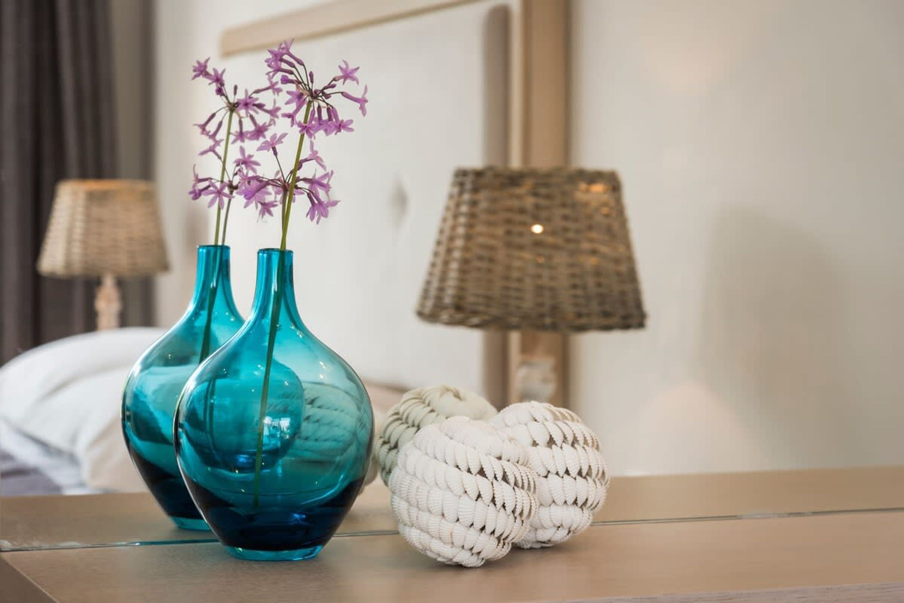 Two teal vases with purple flowers on a bedside table, a woven lamp, and a white textured object beside them.