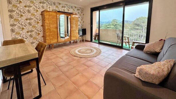 A living room with a sofa, a dining table, and a cabinet. There is a large window with a view of the outdoors.