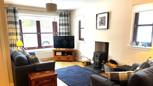 A living room with a sofa, a television, a fireplace, and a window with curtains. - Charming two-bedroom pet friendly property with fire (Aviemore)
