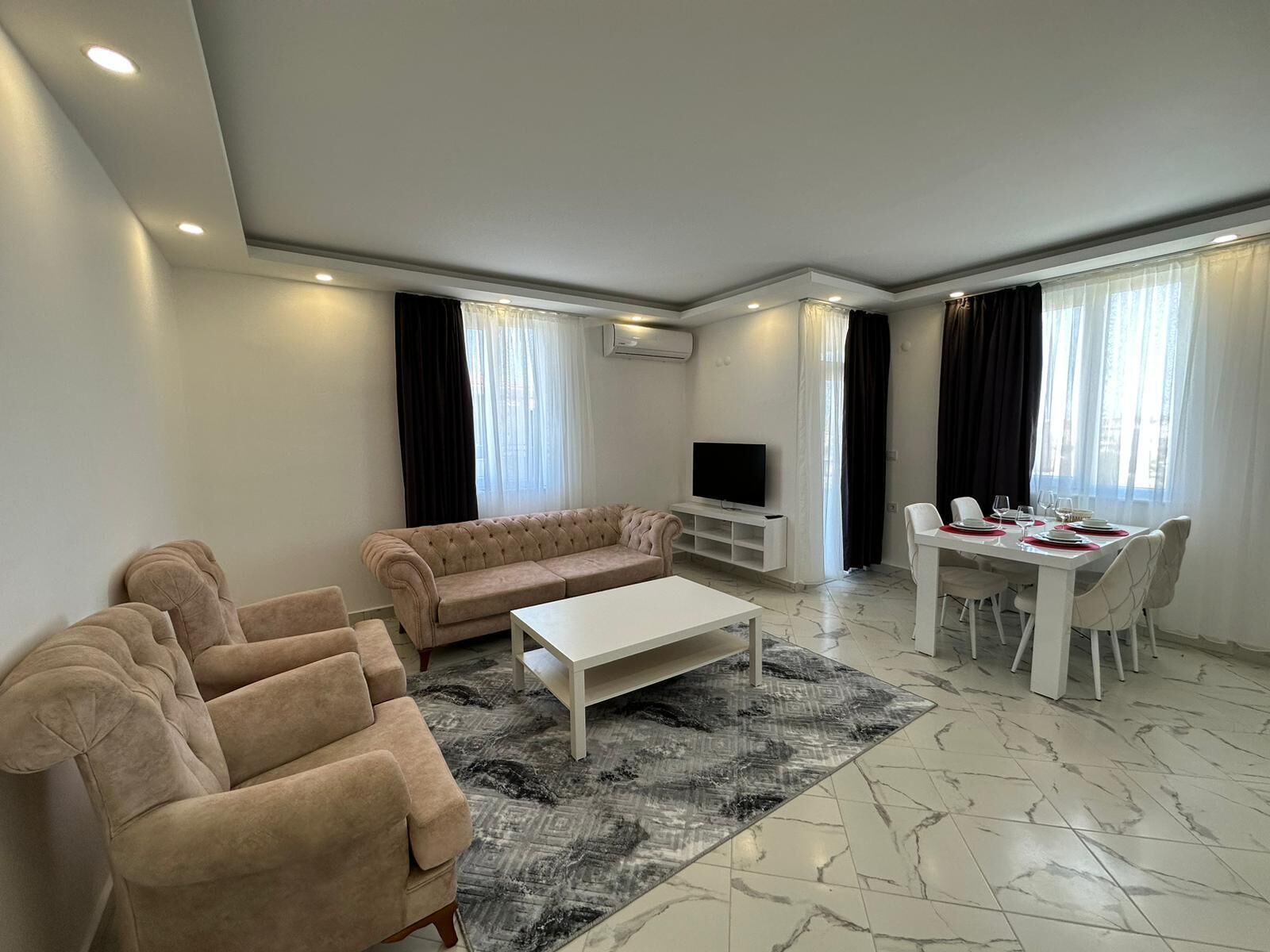 Ein modernes Wohnzimmer mit einem beigefarbenen Sofa, einem weißen Couchtisch, einem Fernseher auf einem Lowboard, einem Esstisch mit Stühlen und einem gemusterten Teppich.
