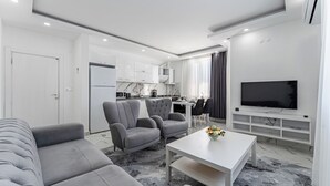 Un salon moderne avec un canapé gris, deux fauteuils, une table basse et un téléviseur posé sur un meuble blanc.
