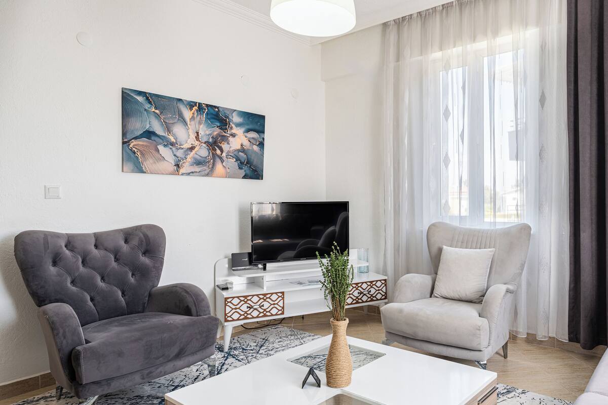 A modern living room with a large flat-screen TV, two armchairs, a coffee table, and a decorative vase.