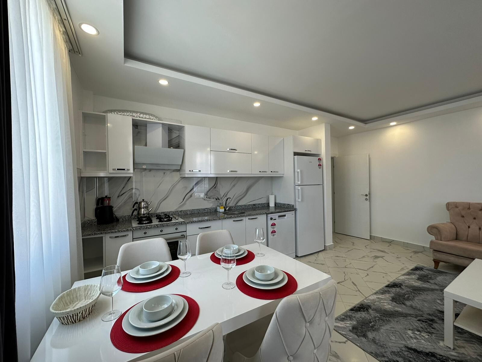 A modern kitchen with white cabinets, a dining table set with plates and glasses, and a beige sofa in the living area.