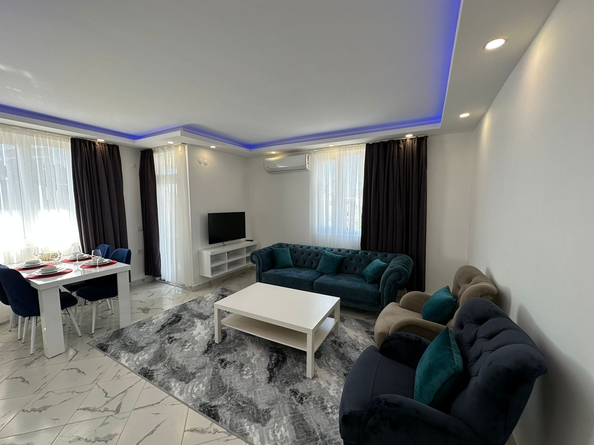 A modern living room with a white TV stand, a teal sofa, a white coffee table, and a dining area with a white table and chairs.