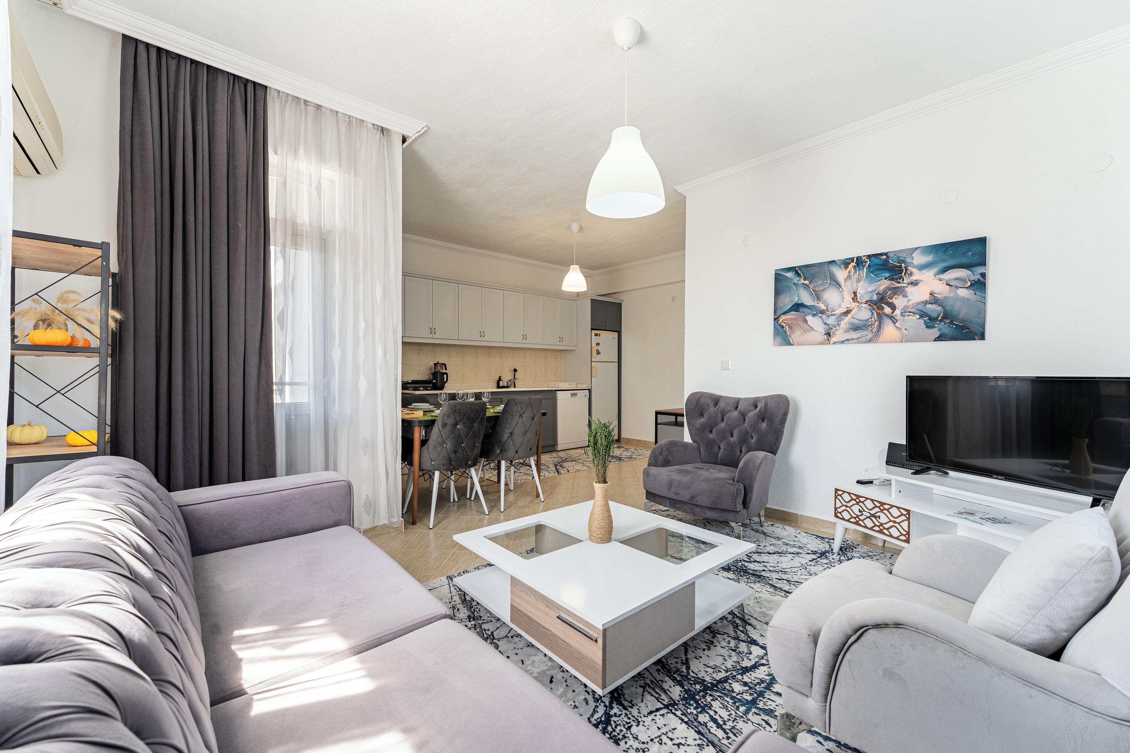 A modern living room with a sofa, a coffee table, and a TV. There is a dining area with chairs and a table, and a kitchen in the background.