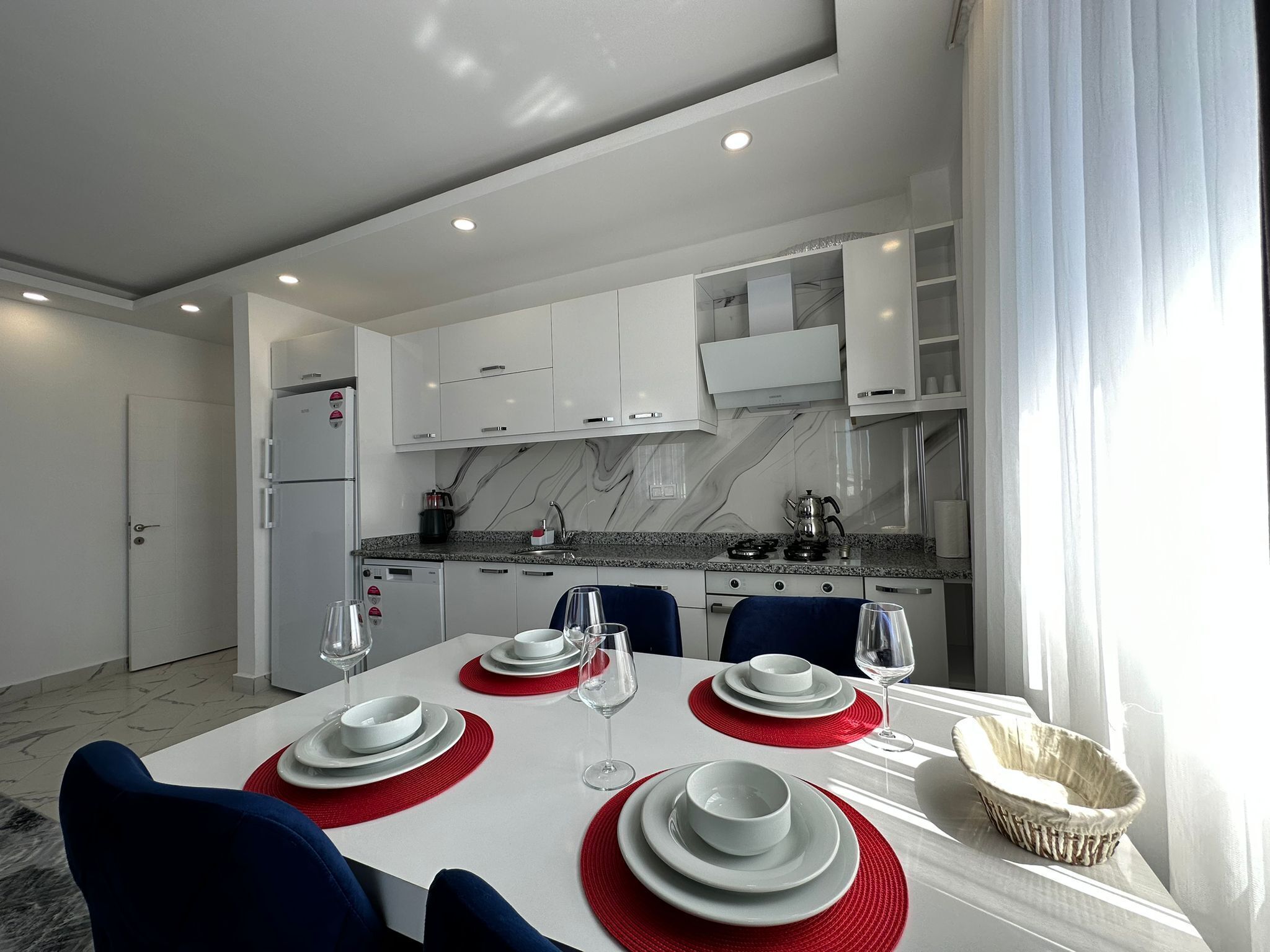 Una cocina moderna con armarios blancos, salpicadero de mármol y un comedor con platos blancos, individuales rojos y sillas azules.
