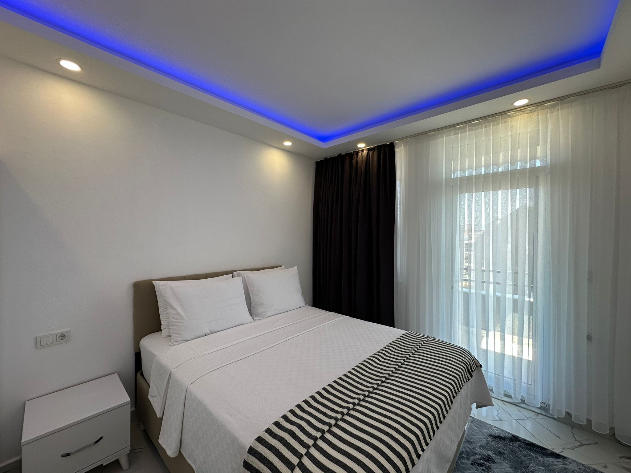 Un dormitorio moderno con cama, mesita de noche y ventana con cortinas translúcidas.