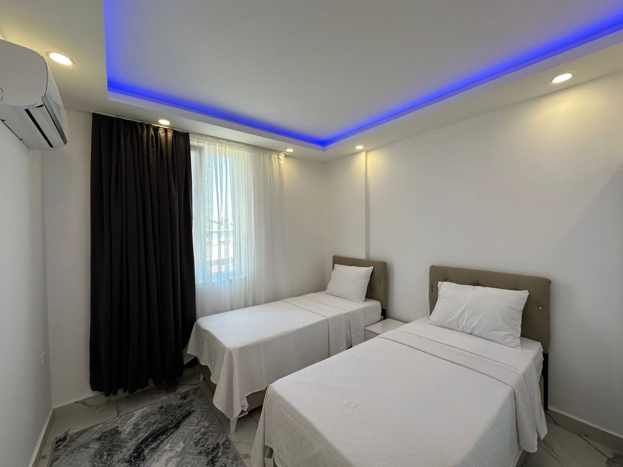 Habitación de hotel con dos camas individuales, un ventanal con cortinas translúcidas, techo con iluminación LED azul y un equipo de aire acondicionado en la pared.