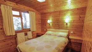A wooden cabin room with a bed, a window with curtains, a nightstand, and a wall-mounted lamp.