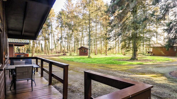 A wooden deck with a table and chairs, overlooking a grassy area with trees and a small cabin.