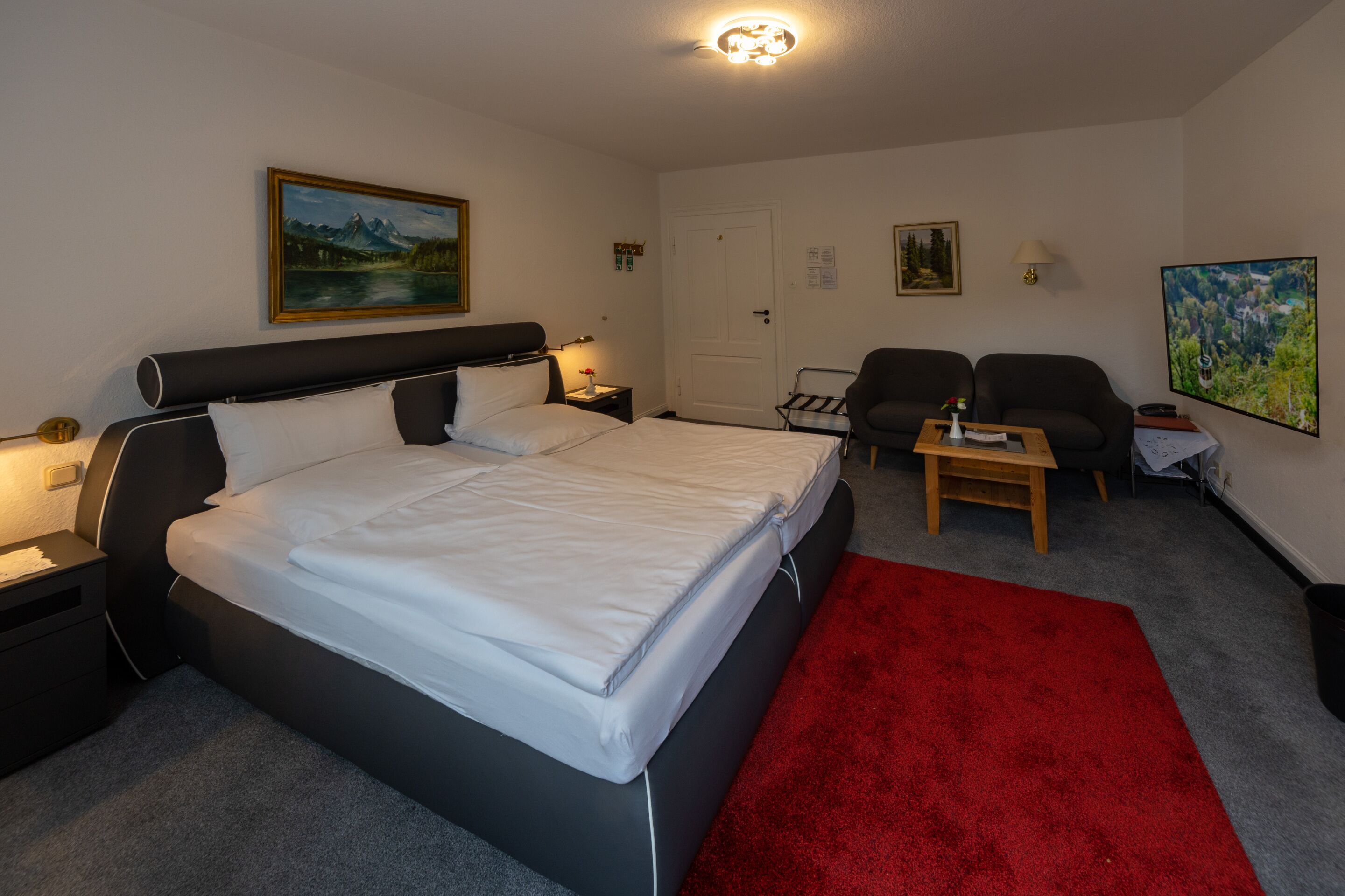 Una habitación de hotel con una cama grande, una alfombra roja, un escritorio, dos sillas y obras de arte en las paredes.