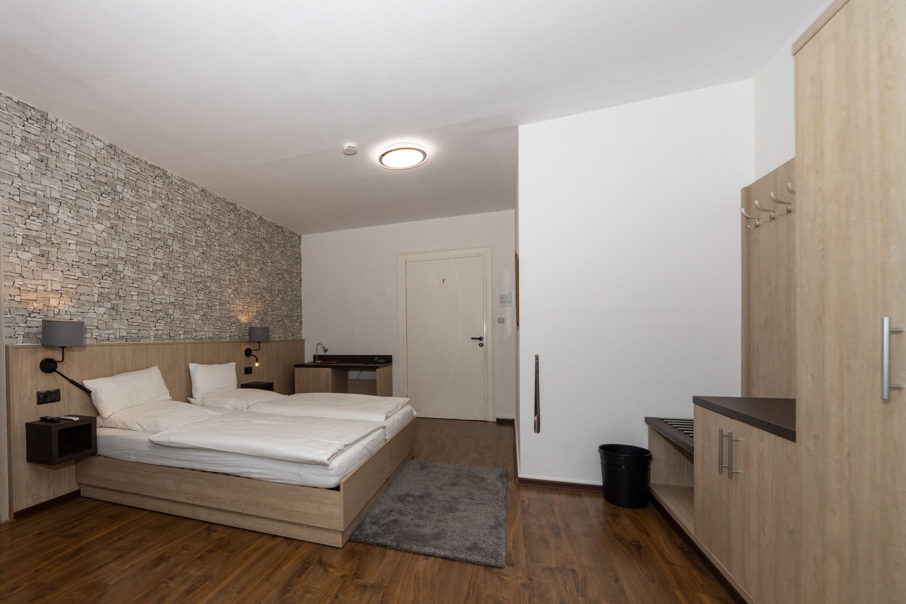 A modern bedroom with a stone wall, a wooden bed, a desk, and a wardrobe.