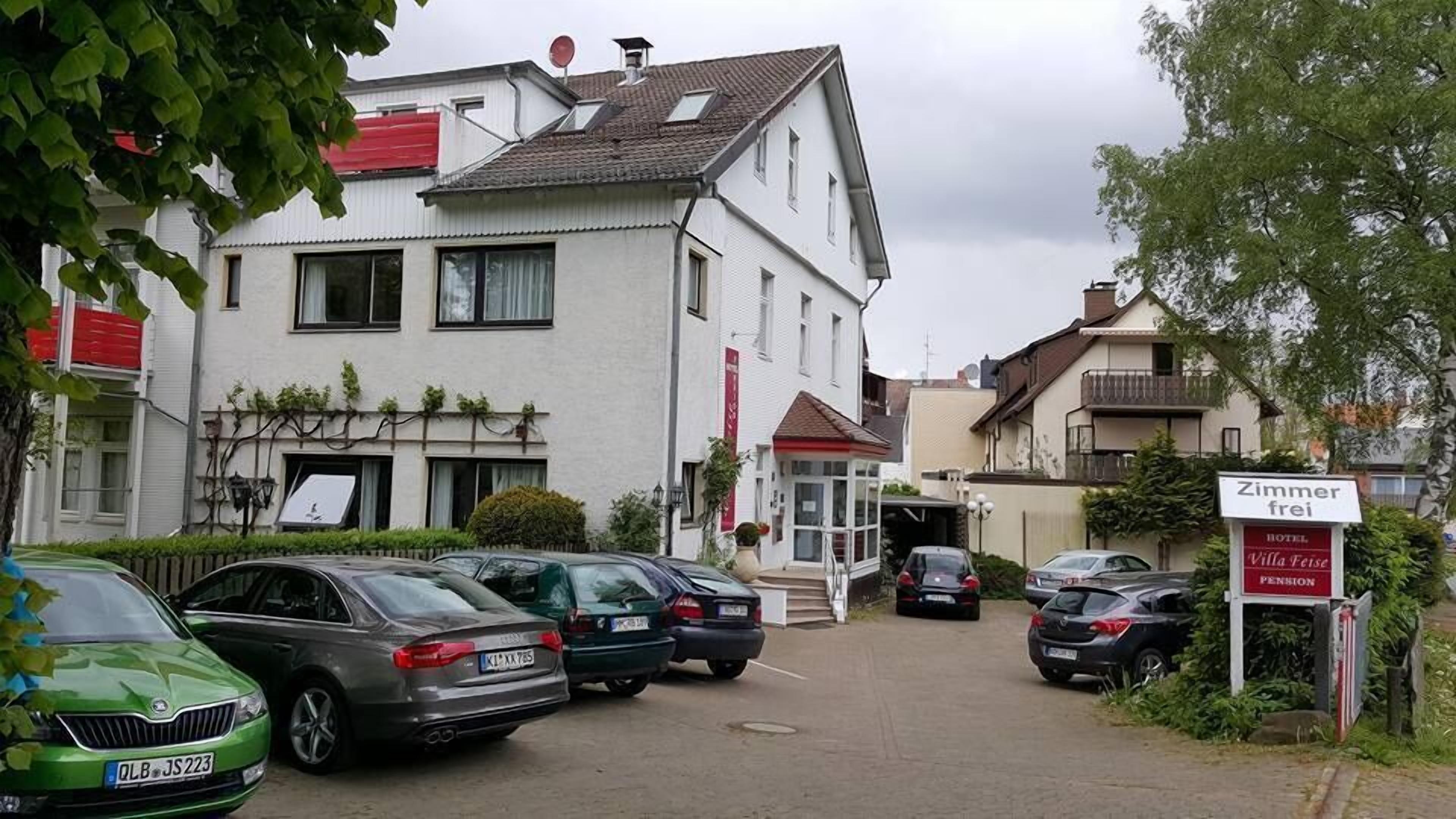 Vista de la calle con coches aparcados, un edificio con un cartel que indica 'Zimmer frei' (habitaciones disponibles) y otro cartel para 'Villa Feise Pension'.