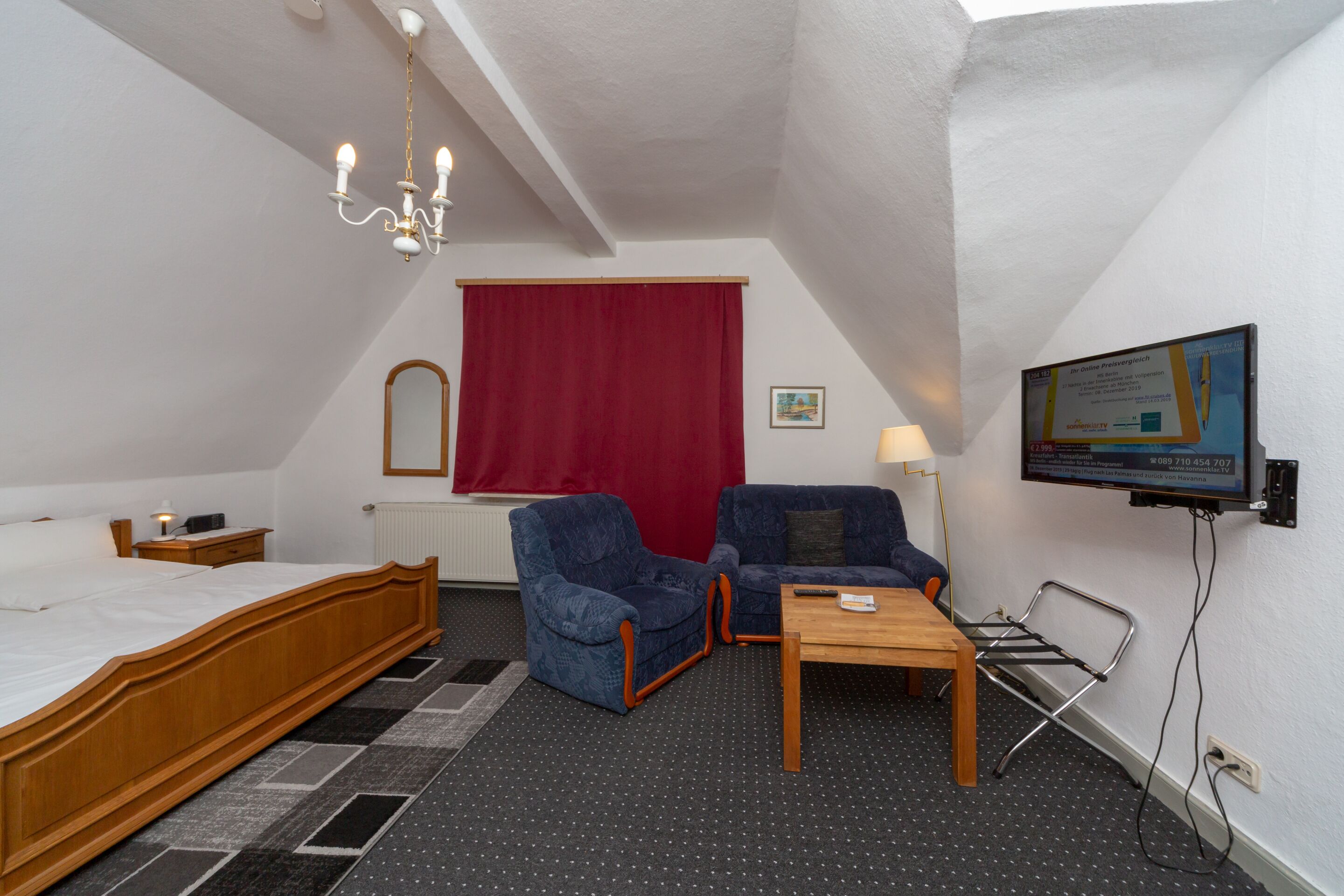 A hotel room with a bed, two armchairs, a small table, a TV, and a red curtain.