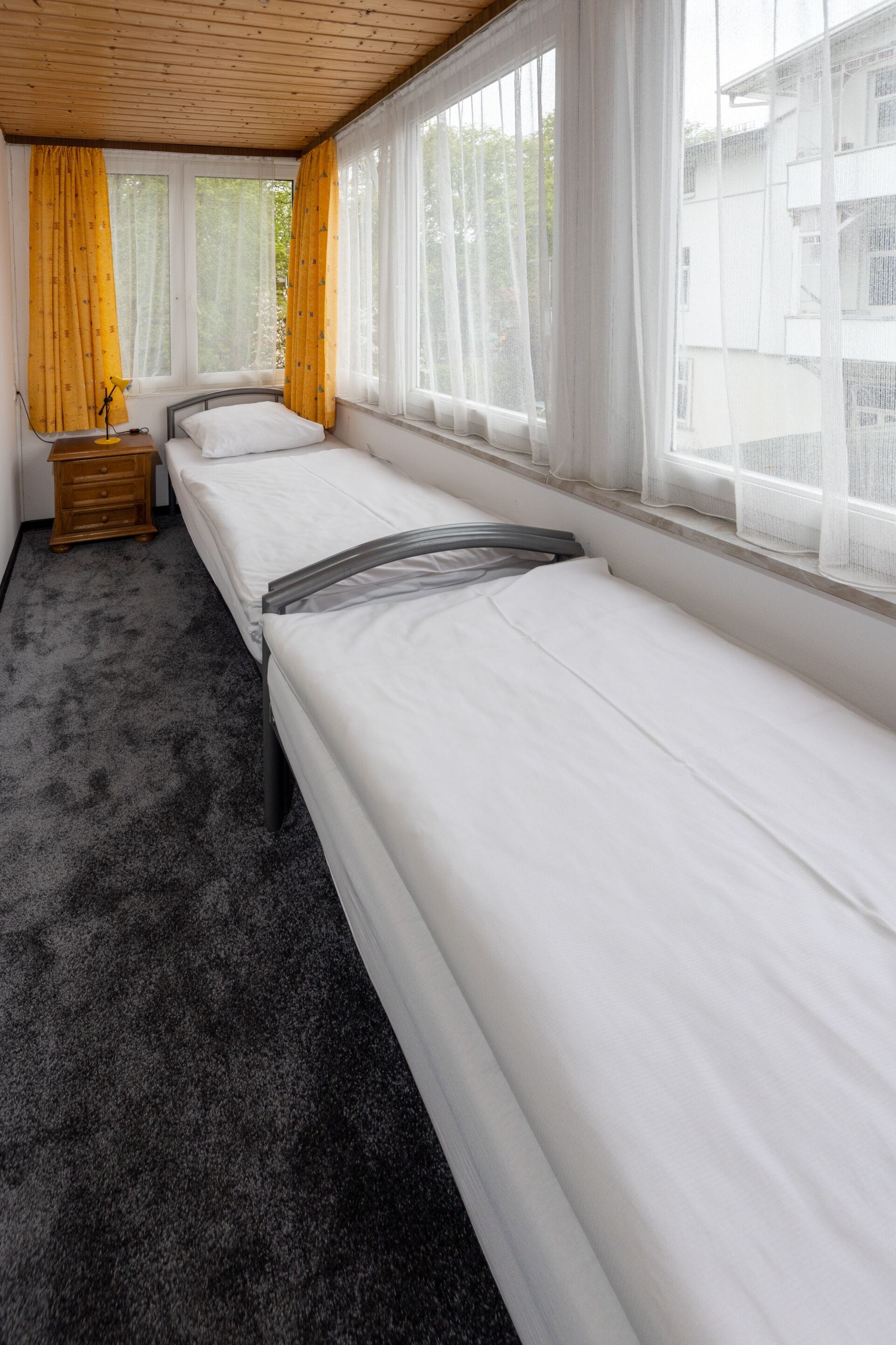 Ein Zimmer mit zwei Einzelbetten, einem Holznachtkästchen, einer Lampe und großen Fenstern mit durchsichtigen Vorhängen.