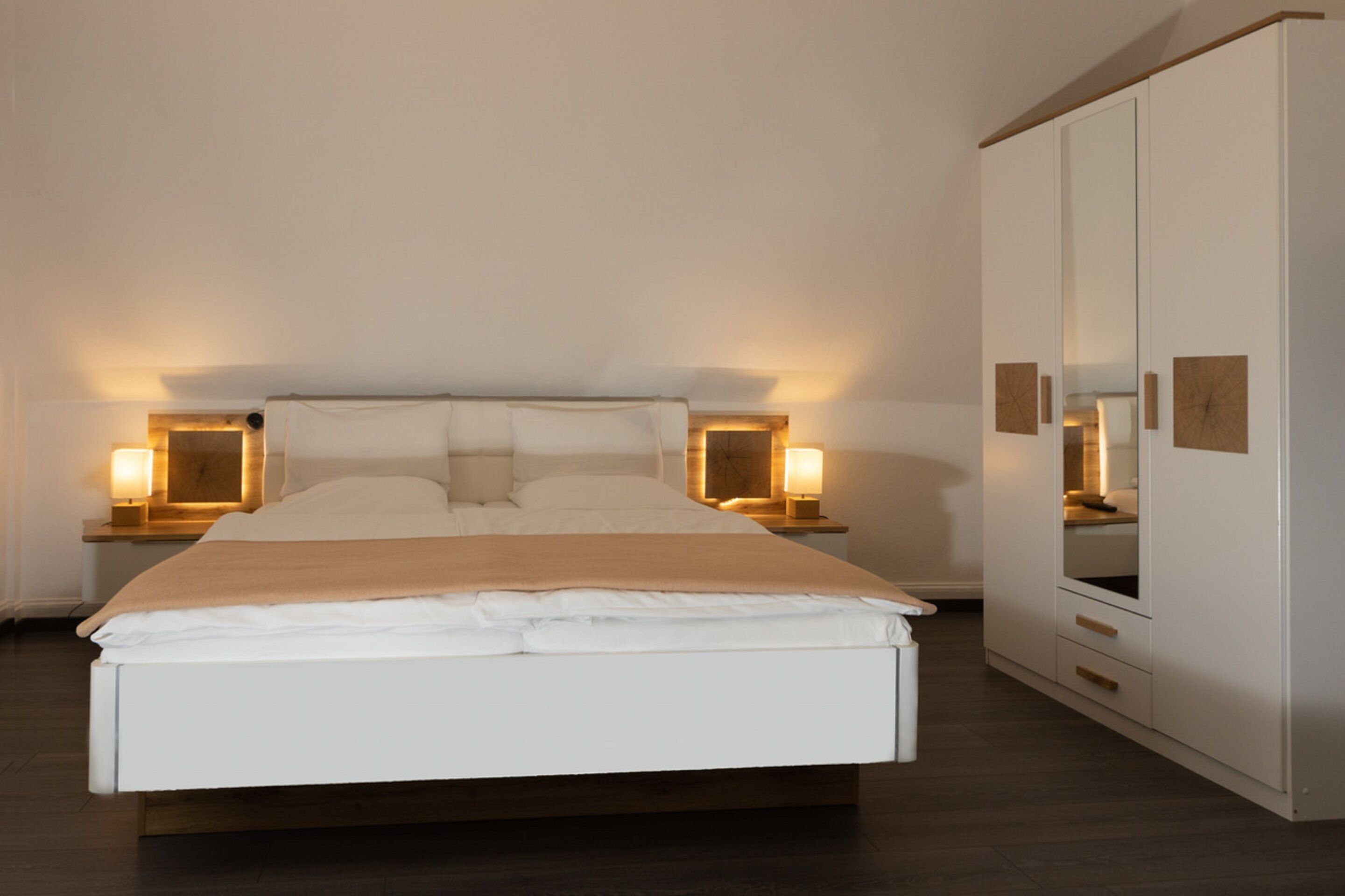 Una cama bien hecha con edredón y almohadones blancos, flanqueada por mesitas de noche con lámparas, en una habitación con un armario grande y un espejo.