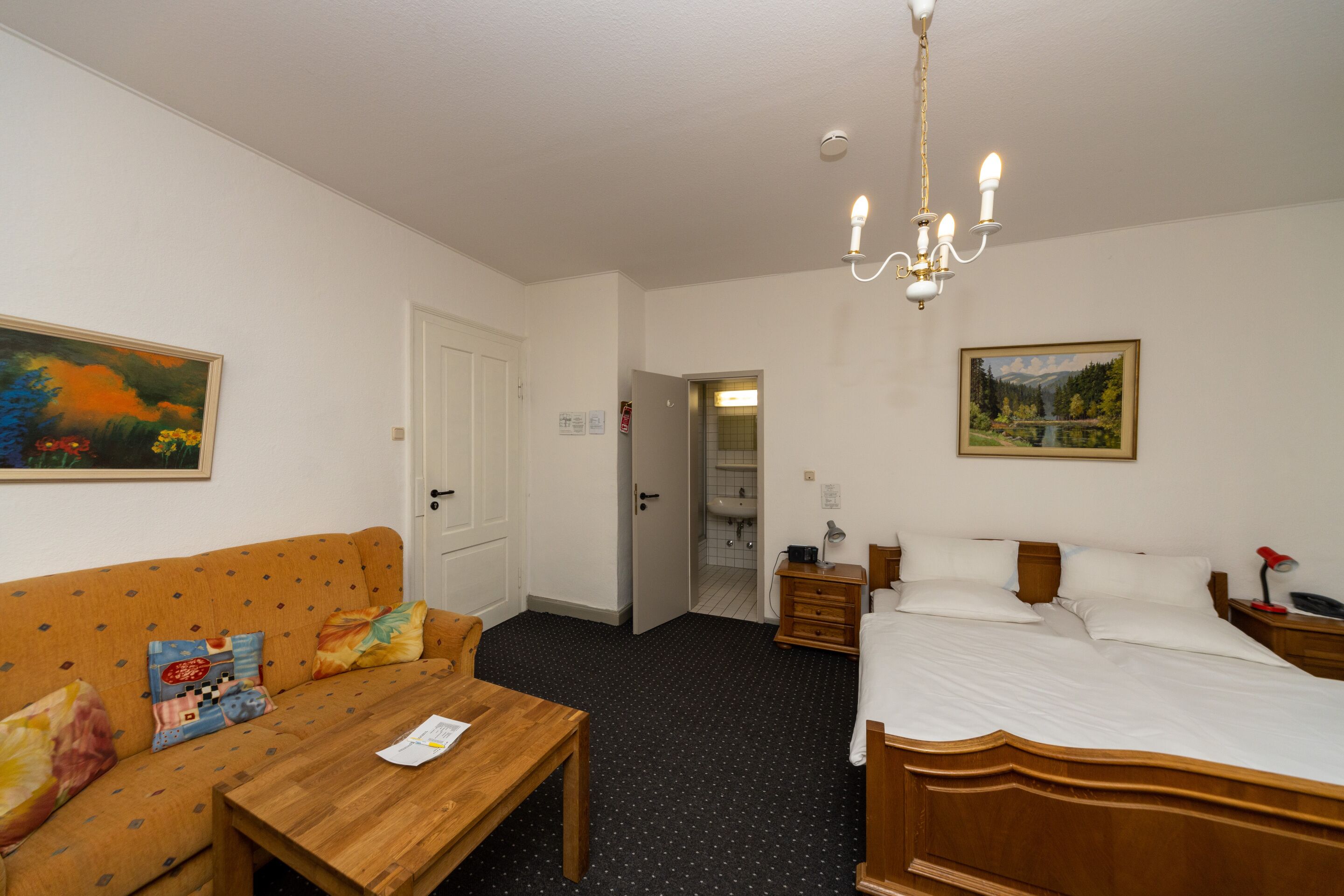 A hotel room with a wooden bed, a wooden table, a sofa with colorful cushions, a chandelier, and a painting on the wall.