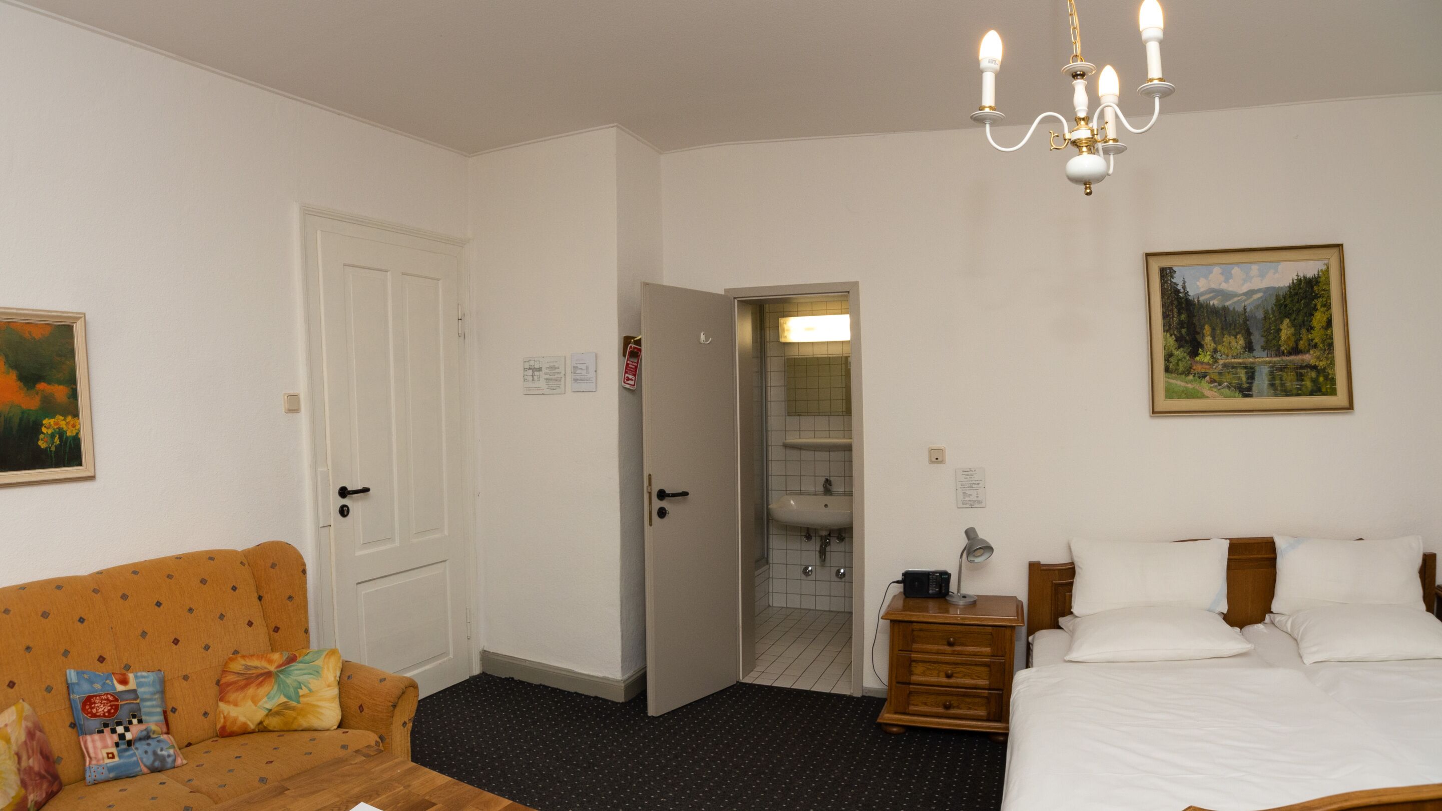 A hotel room with a bed, a wooden nightstand, a lamp, a painting, a door, and a bathroom visible through an open door.