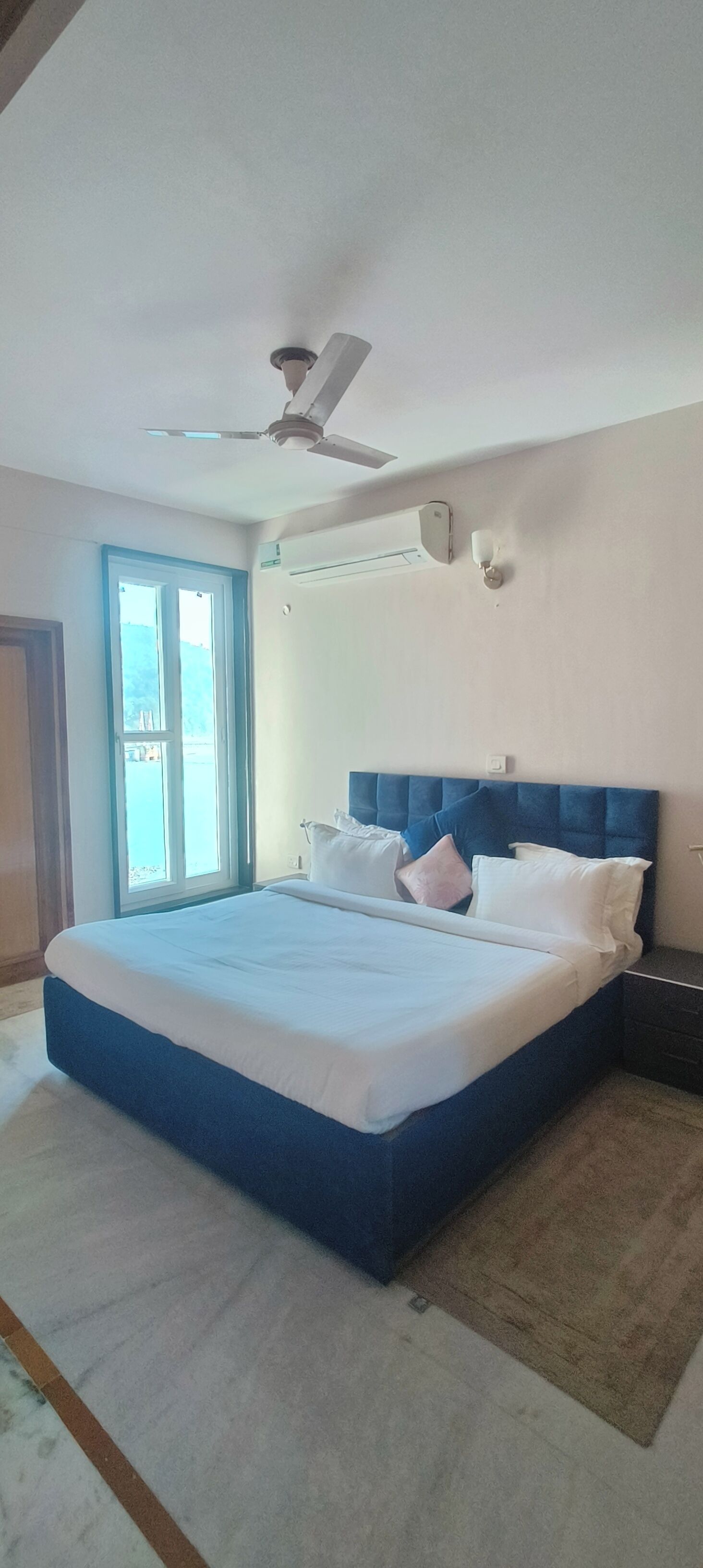 A bedroom with a large bed, a ceiling fan, an air conditioning unit, a window, and a door.
