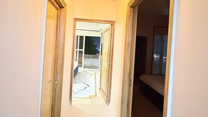 Un couloir étroit, éclairé par un luminaire au plafond, avec des portes en bois et une vue sur une pièce dotée d’une fenêtre.