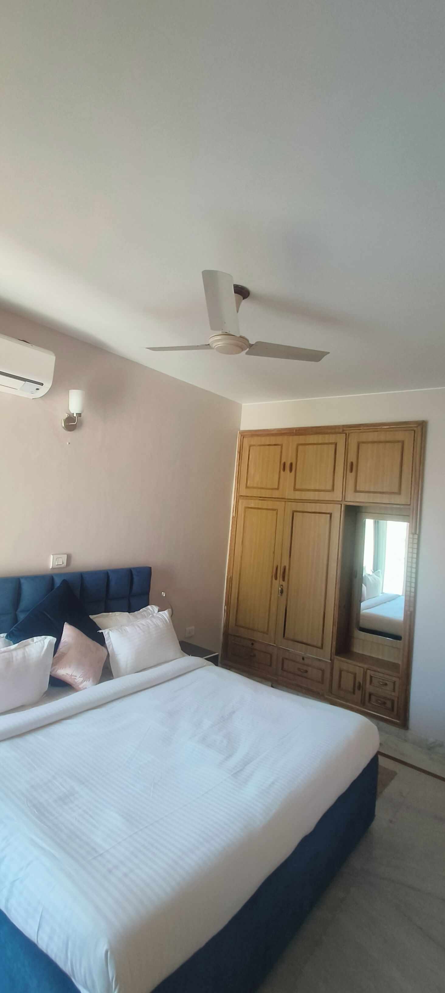 Une chambre avec un grand lit, un ventilateur de plafond, une armoire en bois et une petite fenêtre.