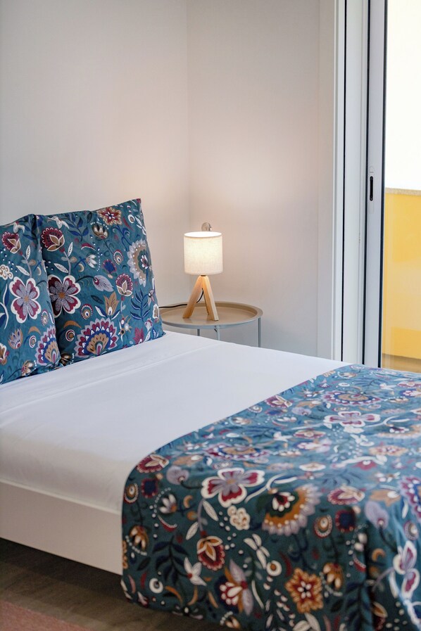 Una cama con colcha y almohadas estampadas con motivos florales, una lámpara de noche y vistas de una pared amarilla a través de una puerta de vidrio.