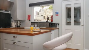 A modern kitchen with a wooden countertop, a coffee maker, a glass of orange juice, and a plate of croissants. There is a white chair and a white door in the background. - 15 Summerland Avenue (Dawlish)