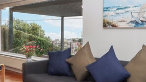 A modern living room with a grey sofa, blue and brown cushions, a large window with a view of greenery and buildings, and a painting of a beach scene. - 15 Summerland Avenue (Dawlish)