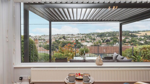 A balcony with a view of a town, a table with a cup and pastries, and a ship decoration. - 15 Summerland Avenue (Dawlish)