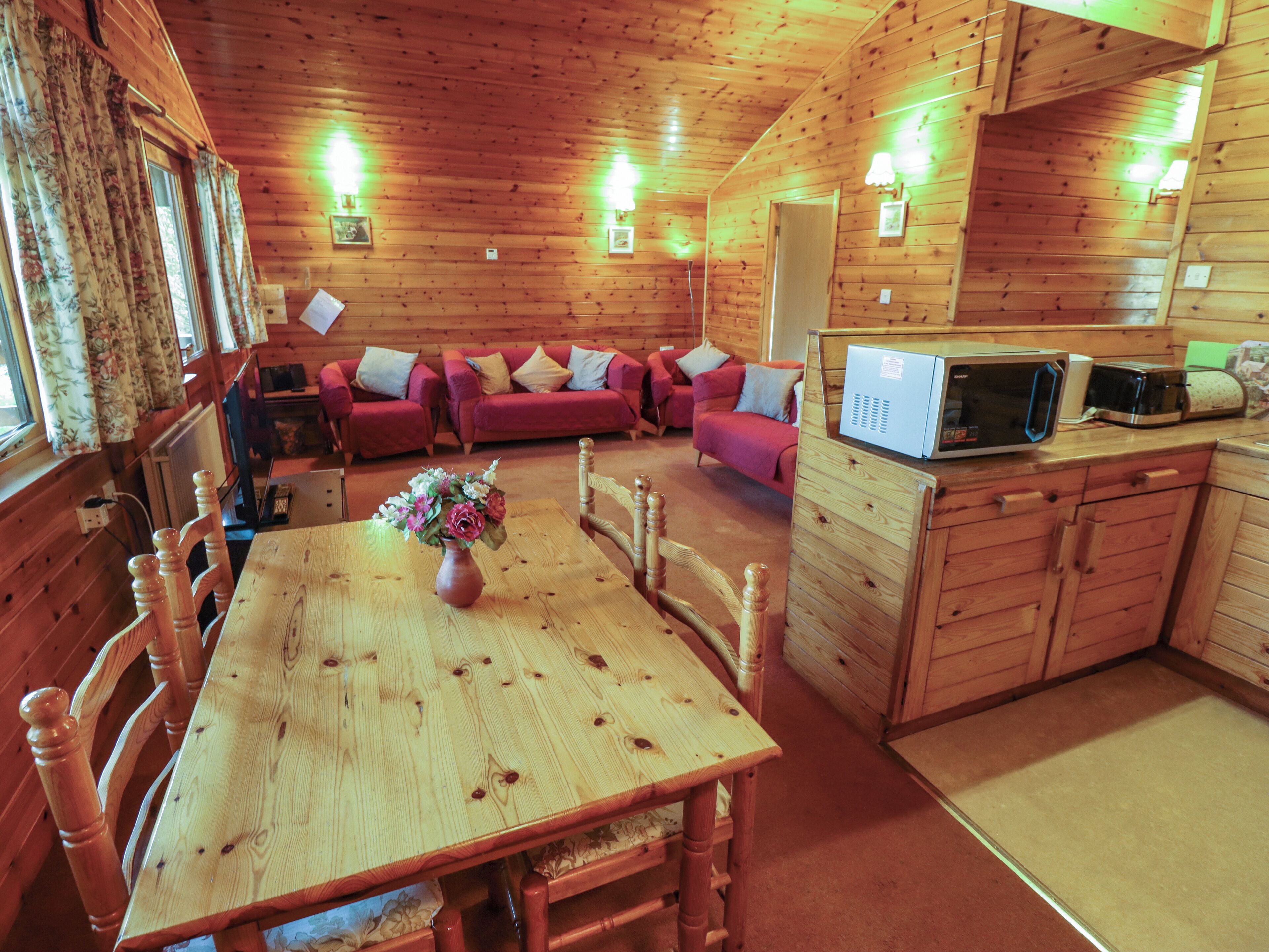 A wooden cabin interior with a dining table, chairs, a sofa set, a microwave, and a small kitchen area.