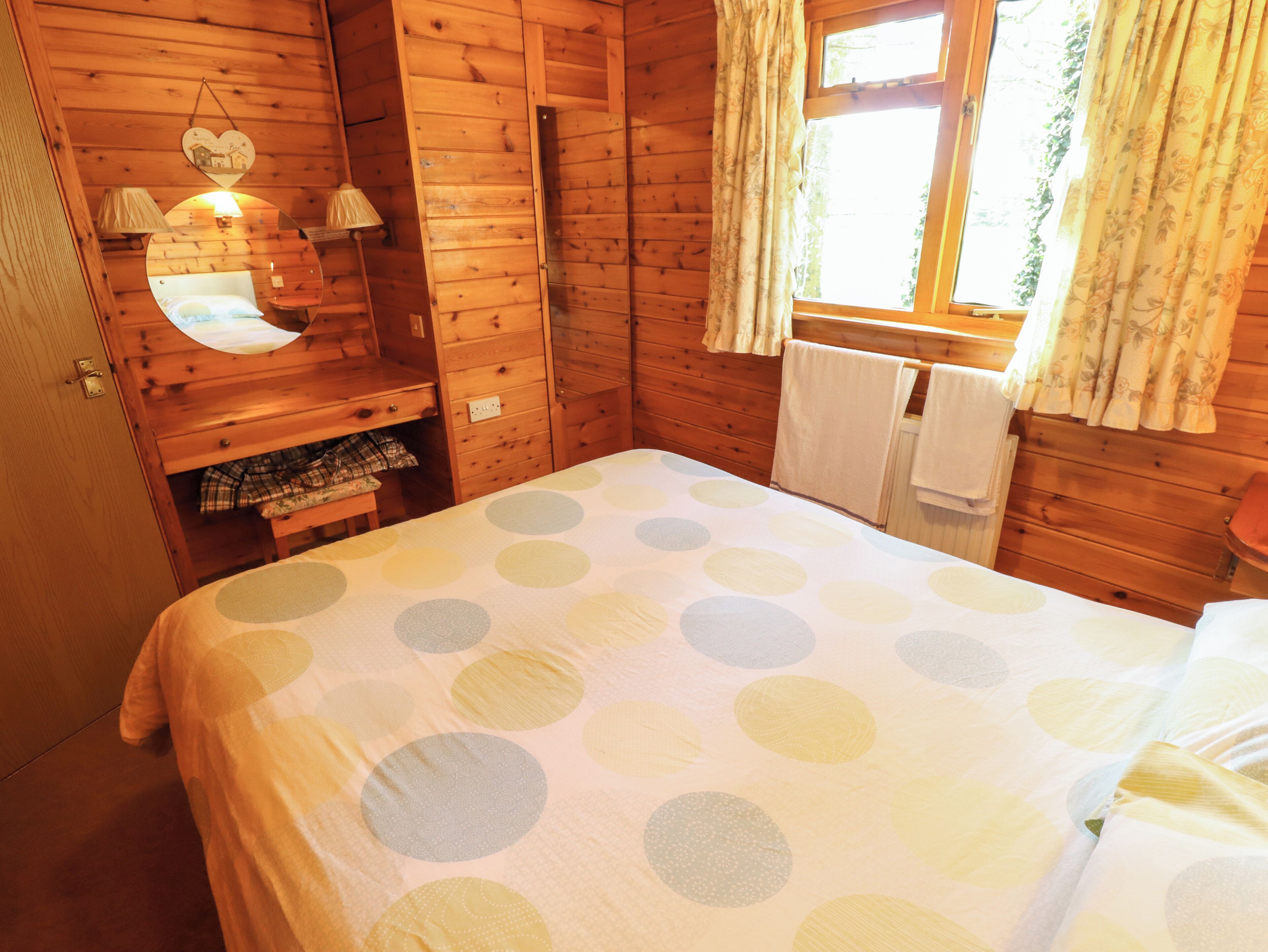A wooden cabin room with a bed, a mirror, a small table, and a window with curtains.