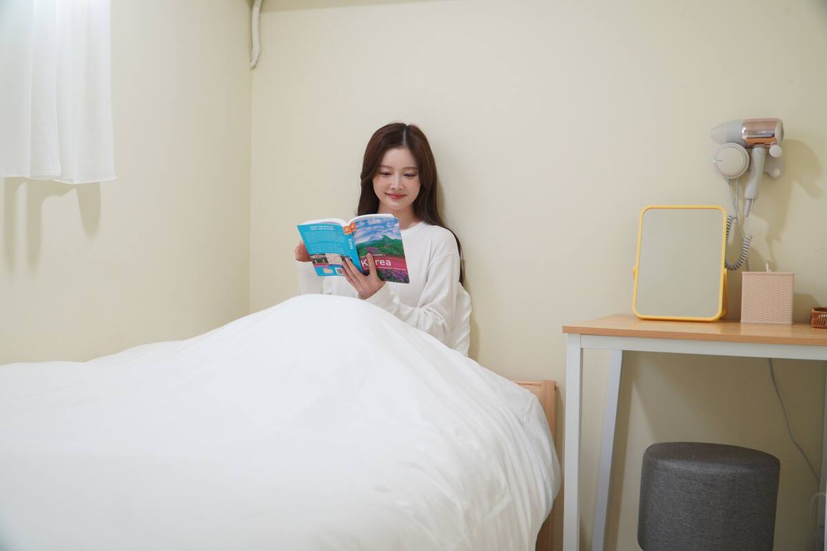 Una persona seduta su un letto che legge un libro in una stanza con un comodino, un asciugacapelli e uno specchio.