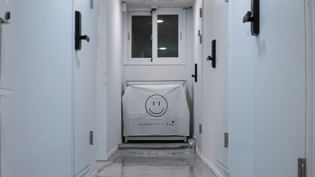 Un pasillo estrecho con puertas blancas a ambos lados, una ventana al final y una bolsa con una cara sonriente y texto en el suelo.