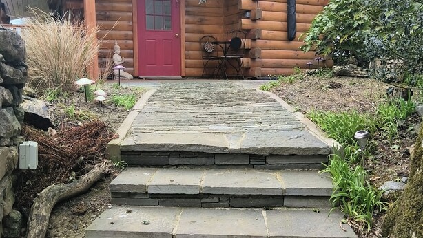 Una cabaña de troncos con puerta roja, escalones de piedra y un jardín con plantas y una estatua.