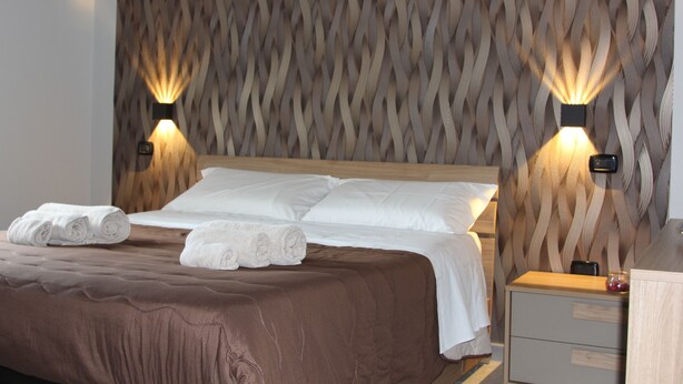 A neatly made bed with white linens and rolled towels, a wooden headboard with a textured pattern, and two wall-mounted lamps on either side. - AGAS Exclusive Apartment (Catanzaro)