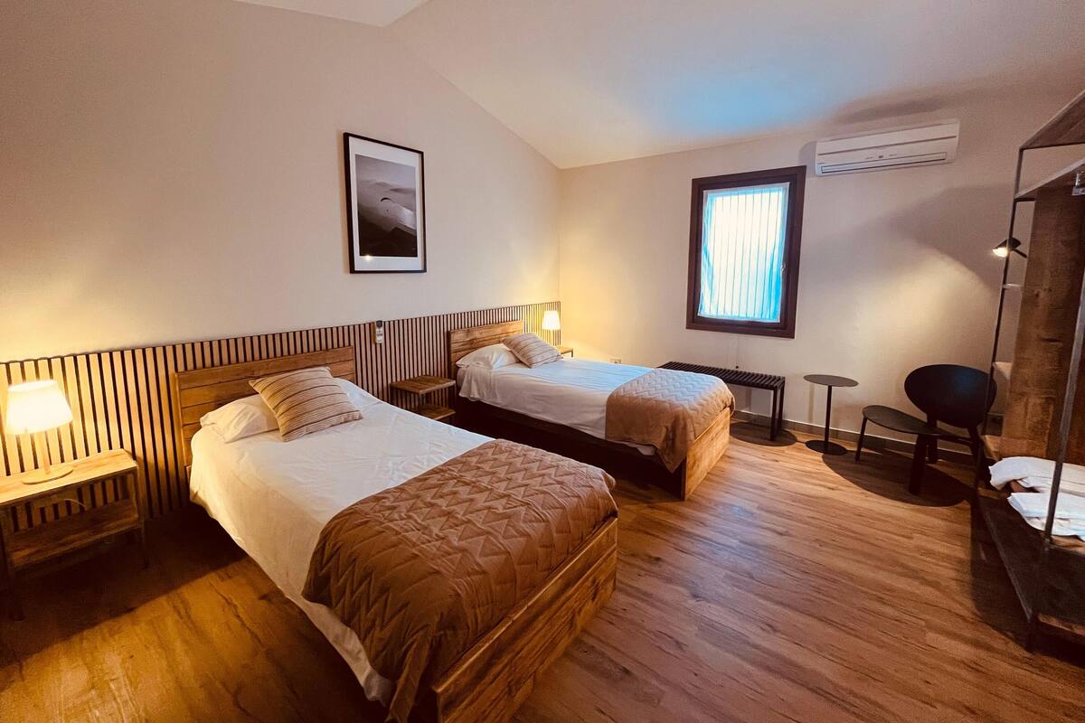 Hotellihuone, jossa on kaksi sänkyä, pieni pöytä, tuoli, ikkuna, jossa on sälekaihtimet, ja ilmastointilaite.