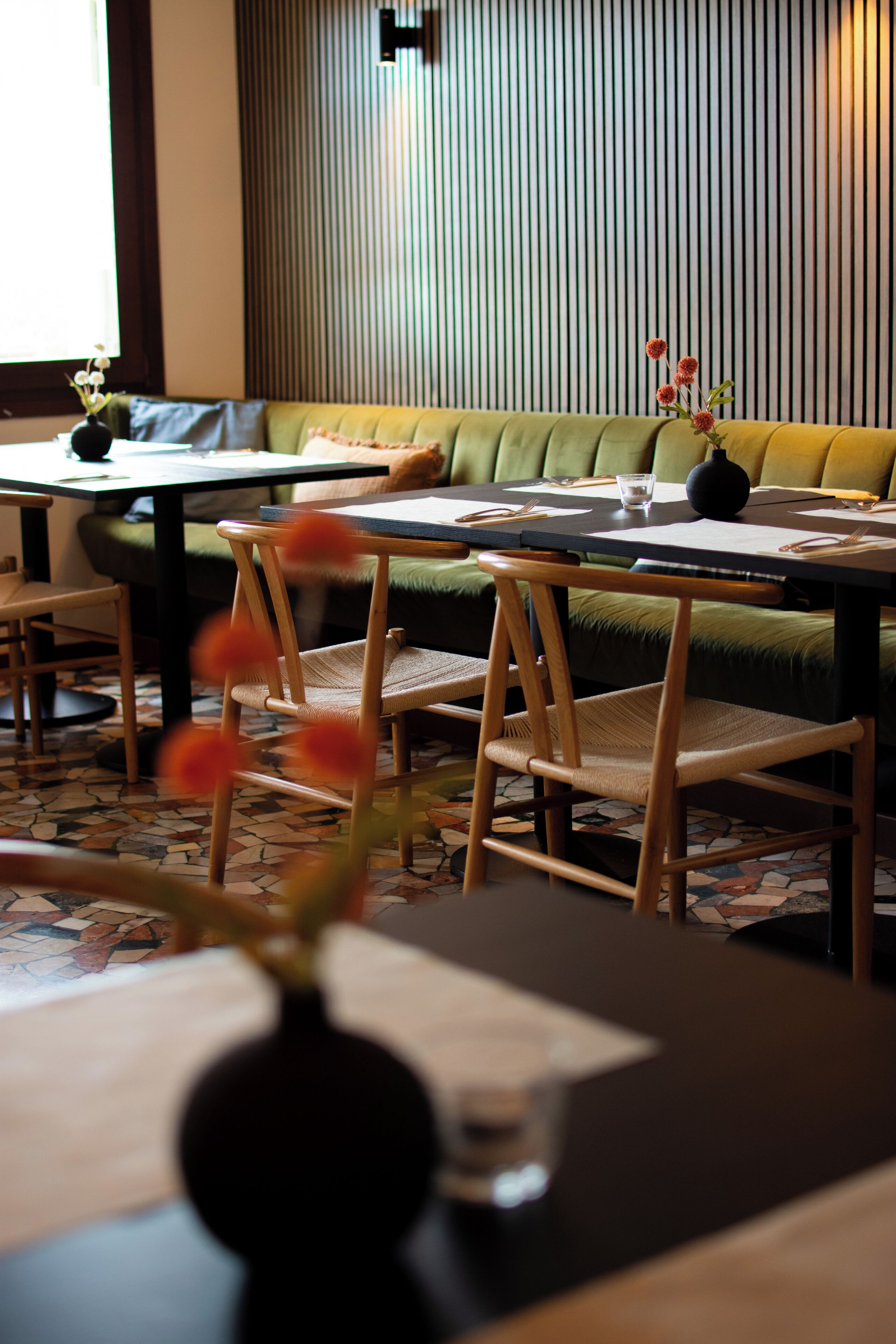 Un interno ristorante con una panca imbottita verde, sedie di legno, un tavolo con un vaso di fiori e un pavimento decorato.