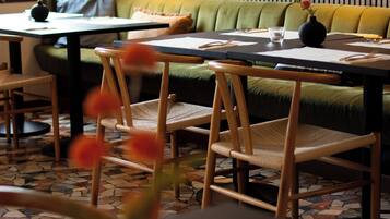 Interior de restaurante con un banco acolchado verde, sillas de madera, una mesa con un florero y suelo estampado.