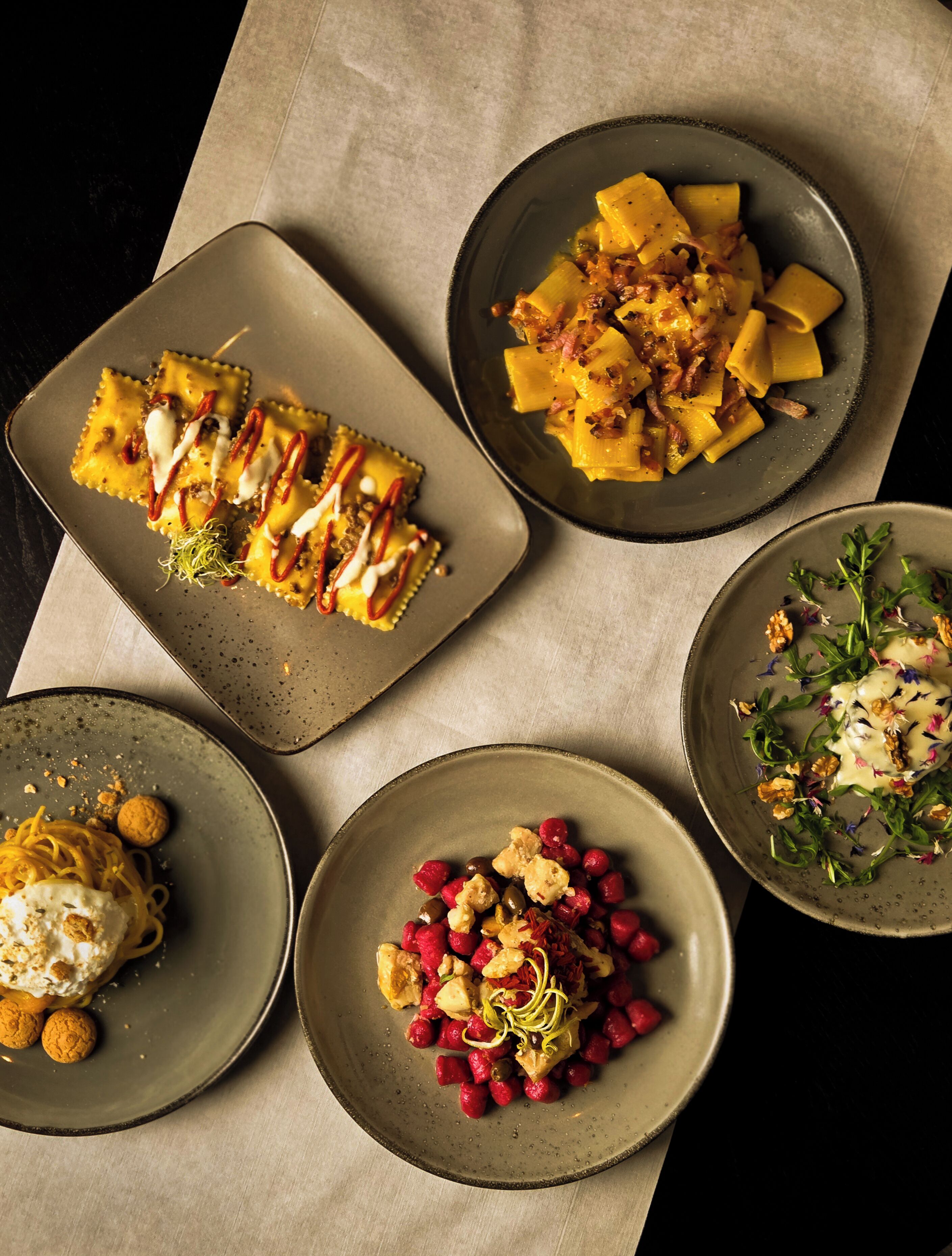 Variedad de platos, incluyendo ravioles, pasta con queso y tocino, y una ensalada con nueces y bayas.