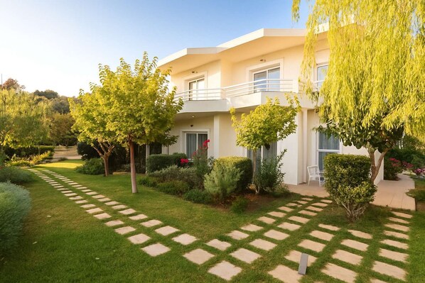 A modern two-story building with a well-maintained garden, paved pathways, and a variety of trees and shrubs.