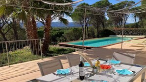 An outdoor dining area with a table set for two, a bottle of wine, and a view of a swimming pool and mountains in the background.