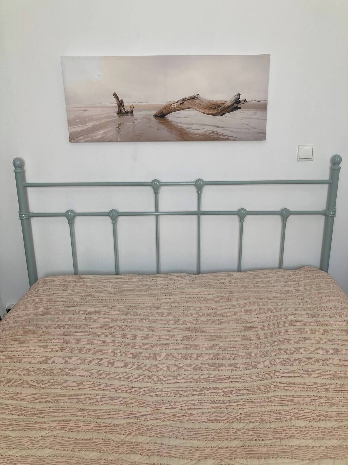 A bed with a patterned bedspread, a metal headboard, and a painting of a beach scene on the wall.