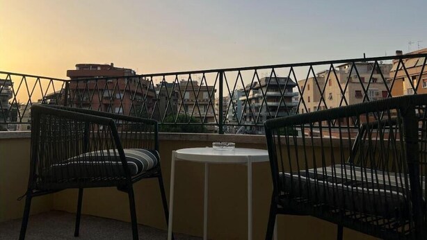 A balcony with two chairs and a small table, overlooking a cityscape during sunset.