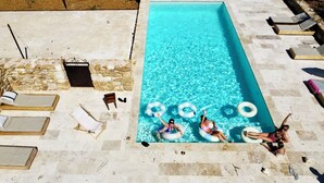 Une piscine rectangulaire à l’eau d’un bleu limpide, entourée de murs en pierre et dotée d’un sol carrelé. On y trouve des chaises longues et une petite table en bois près de la piscine.