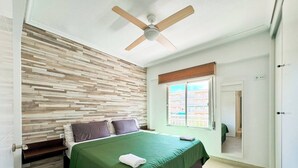 Un dormitorio con cabecera de madera, ventilador de techo, ventana con vistas a edificios y colcha verde.