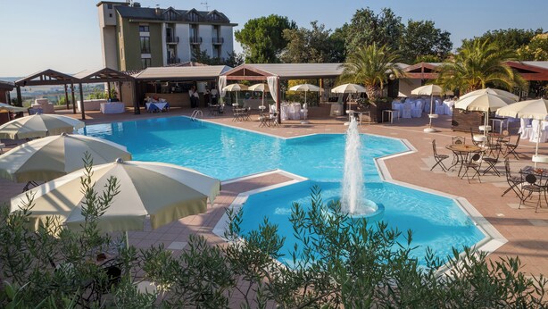 A pool area with multiple lounge chairs and umbrellas, a fountain, and a building in the background. - Hotel La Cantinella (Ostra)