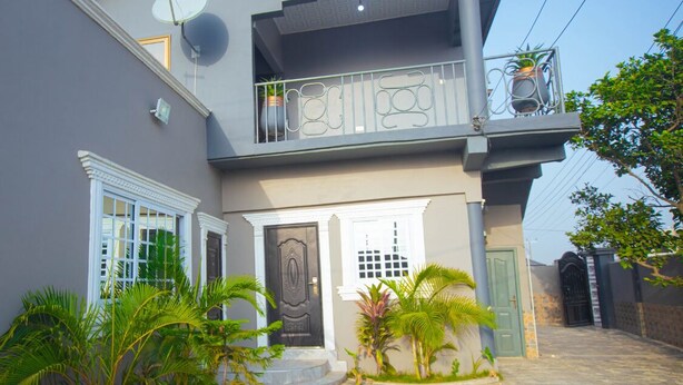 A two-story house with a balcony, satellite dish, and a gated entrance. - GHL. G Floor En-suites 2BR Sleep 4. WiFi, DSTV, SOLAR ,AC And Free Breakfast (Kasoa Ghana)